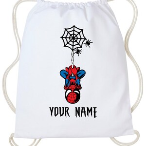 Puede incluir: Mochila con cord&oacute;n blanca con un dise&ntilde;o de Spiderman. El dise&ntilde;o incluye una telara&ntilde;a negra, dos peque&ntilde;as ara&ntilde;as y Spiderman colgado boca abajo. El texto "YOUR NAME" est&aacute; impreso en negro debajo.