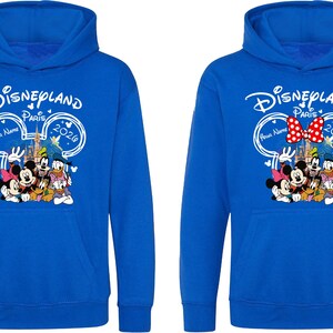 Puede incluir: Dos sudaderas azules con un gr&aacute;fico de Disneyland Paris 2020. El gr&aacute;fico presenta a Mickey y Minnie Mouse, el Pato Donald y Goofy. Una sudadera tiene un lazo rojo y blanco. La otra tiene un espacio en blanco para un nombre.