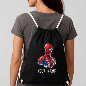 May include: Black drawstring backpack featuring a vibrant Spider-Man graphic. The superhero is depicted in red and blue, with a white spider emblem. The text "YOUR NAME" is printed in white below the image. White cords serve as straps.