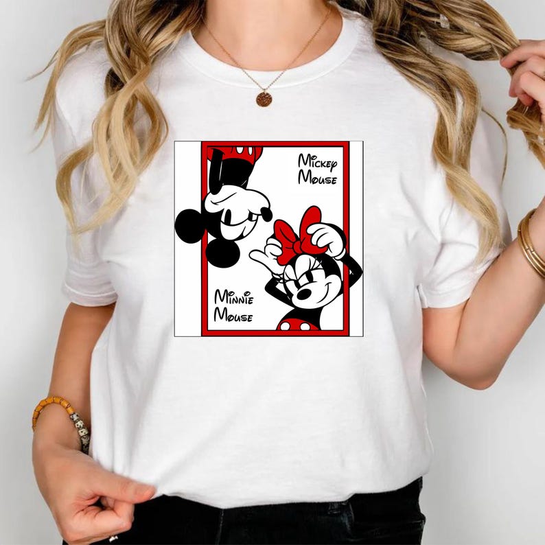 May include: White t-shirt featuring a framed graphic of Mickey and Minnie Mouse. Mickey is upside down, and Minnie is smiling with a red bow. The words "Mickey Mouse" and "Minnie Mouse" are printed on the graphic.