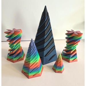 May include: A collection of five abstract geometric sculptures. Two are multi-colored, with a spiral design. One is dark blue with black stripes. The other two are smaller, with a rainbow color scheme and a ridged design. The sculptures are arranged on a white surface.