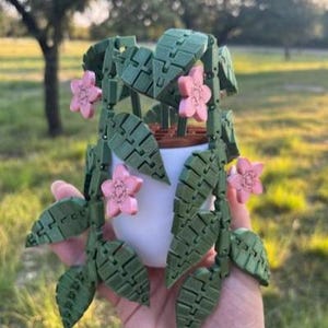 May include: A white ceramic pot with a green, articulated plant and pink flowers. The plant's leaves and flowers are detailed, creating a unique, decorative piece. The plant is held in a hand, with a blurred outdoor background.