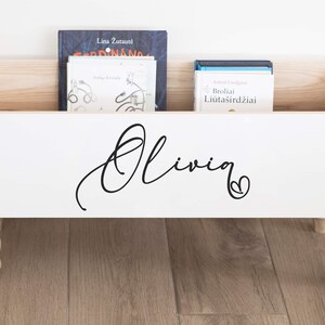 May include: White wooden storage box with a personalized name "Olivia" in black cursive lettering and a small heart. The box has four turned wooden legs and is filled with books.