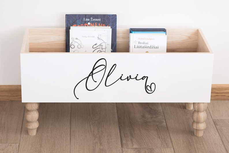 May include: White wooden storage box with a personalized name "Olivia" in black cursive lettering and a small heart. The box has four turned wooden legs and is filled with books.