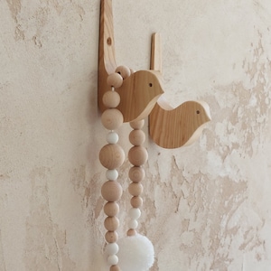 Puede incluir: Dos ganchos de pared de madera con forma de p&aacute;jaro con una guirnalda de cuentas colgando de ellos. La guirnalda est&aacute; hecha de cuentas de madera natural y pompones blancos.