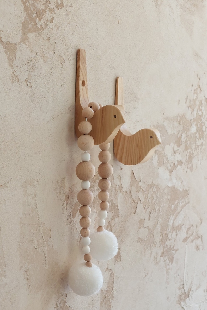 Puede incluir: Dos ganchos de pared de madera con forma de p&aacute;jaro con una guirnalda de cuentas colgando de ellos. La guirnalda est&aacute; hecha de cuentas de madera natural y pompones blancos.