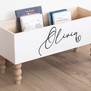 May include: White wooden storage box with turned legs and a black inscription that reads "Olivia" with a heart. The box is filled with books.