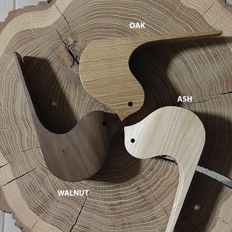 Puede incluir: Tres ganchos de pared de madera con forma de p&aacute;jaro en diferentes tipos de madera: nogal, fresno y roble. Los ganchos est&aacute;n dispuestos en c&iacute;rculo sobre una rodaja de tronco de &aacute;rbol.