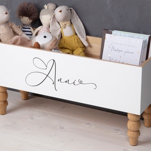 May include: White toy storage box with wooden legs and the name "Annie" written on the front. The box is filled with stuffed animals.