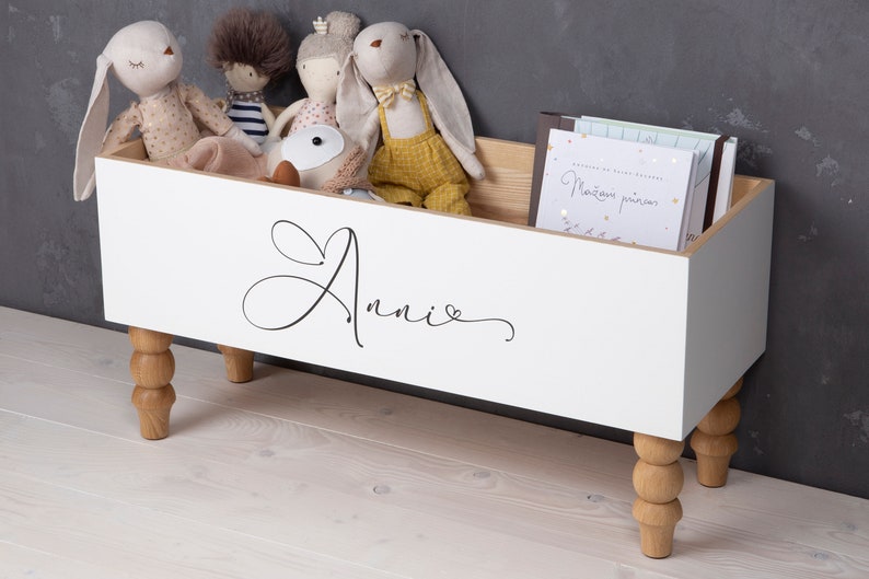 May include: White toy storage box with wooden legs and the name "Annie" written on the front. The box is filled with stuffed animals.