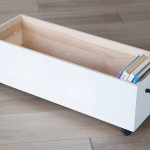 May include: White wooden storage box with black and white bead handle and wheels. The box is open and contains a stack of books.