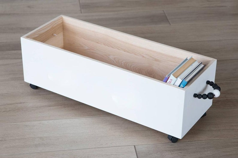 May include: White wooden storage box with black and white bead handle and wheels. The box is open and contains a stack of books.