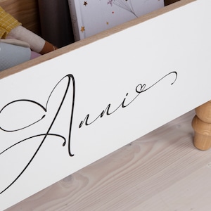 May include: White wooden storage box with a cursive inscription that reads "Annie" with a small heart symbol. The box has wooden legs.