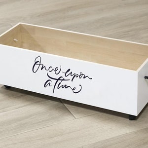 K&ouml;nnte beinhalten: Wei&szlig;e Holzkiste mit schwarzen Rollen und einem schwarzen Perlenhenkel. Die Kiste tr&auml;gt die Aufschrift "Once upon a time" in schwarzer Schrift.