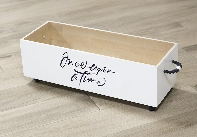 K&ouml;nnte beinhalten: Wei&szlig;e Holzkiste mit schwarzen Rollen und einem schwarzen Perlenhenkel. Die Kiste tr&auml;gt die Aufschrift "Once upon a time" in schwarzer Schrift.
