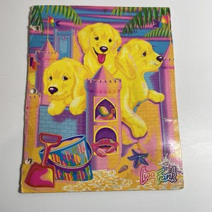 May include: A Lisa Frank-branded notebook cover featuring three golden retriever puppies in front of a sandcastle. The castle is pink and yellow with a blue sky background. A bucket, shovel, and starfish are in the foreground.