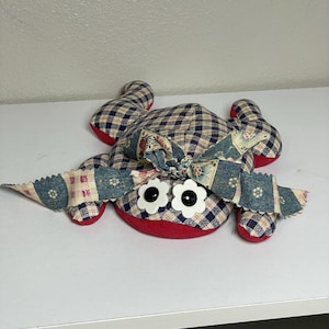 May include: A handmade fabric frog with a red and blue plaid body, red accents, and large white flower-shaped eyes. The frog has a bow on its head and is made of various patterned fabrics.