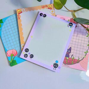 May include: Four colourful memo pads with unique designs. One pad has a blue grid pattern with a pink flower, another has an orange border with bacon and egg illustrations, a third has a purple border with soot sprite characters, and the last has a pink grid with a cat illustration.