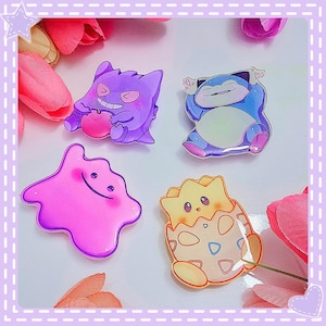 May include: Four character pins in vibrant colours. A purple Gengar with a pink fruit, a blue Snorlax, a pink Ditto, and a yellow Togepi hatching from an egg. The pins are set against a backdrop of pink and purple flowers.