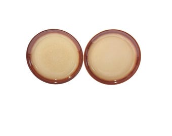 May include: Two round ceramic plates with a light beige center and a dark brown rim. The plates are displayed on a white background. The plates are likely for serving food.