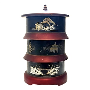 May include: A tiered, cylindrical container with a dark blue finish and red accents. The container features gold and white Asian-inspired landscape designs. The base and top are dark red, and the container has a small, decorative finial.