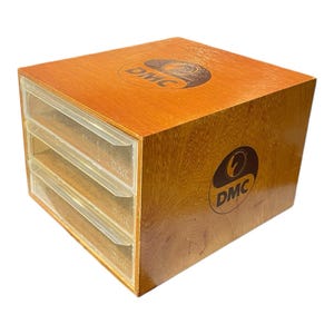 May include: A wooden storage box with three clear plastic drawers. The box is a warm brown color with the DMC logo printed on the top and side. The drawers are labeled with the DMC logo.