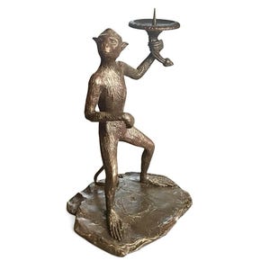 May include: Bronze monkey figurine holding a candle holder. The monkey is standing on a textured base. The candle holder is round with a spike in the center. The figurine is a decorative item.