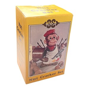 May include: A yellow box with the words "Nut Cracker Set" printed on it. The box features a ceramic monkey nutcracker wearing a red beret and holding paintbrushes and a palette. The monkey is wearing glasses and an apron.