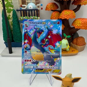 May include: A Charizard ex Pokémon trading card, featuring a blue and red dragon-like creature. The card is displayed on a clear stand, with a small Eevee figure in the foreground. The background includes a miniature autumn-themed display.
