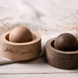 May include: Two wooden ring holders, one light and one dark, each with a round top. The light holder has "john & sarah" engraved with a heart. The dark holder has "HENRY & SARAH EST 2026" engraved. Both are on a white surface.