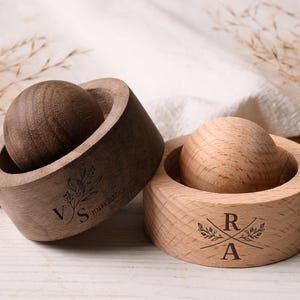 Puede incluir: Dos portaanillos de madera, uno marrón oscuro y otro marrón claro, cada uno con una parte superior esférica. El soporte marrón oscuro tiene las iniciales "VS" y la fecha "10.04.2023" grabadas. El soporte marrón claro tiene las iniciales "RA".