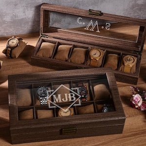 Puede incluir: Dos cajas de madera para relojes, una abierta y otra cerrada, con tapas de cristal. La caja abierta muestra relojes sobre cojines marrones. La caja cerrada tiene las iniciales "MJB" grabadas en el cristal. Tambi&eacute;n se ven un tocadiscos vintage y un reloj sobre una almohada.