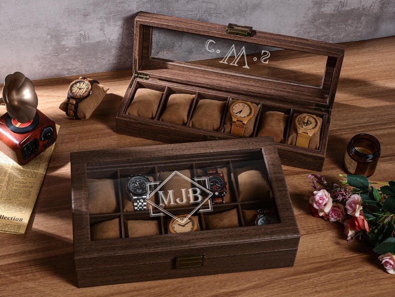 Puede incluir: Dos cajas de madera para relojes, una abierta y otra cerrada, con tapas de cristal. La caja abierta muestra relojes sobre cojines marrones. La caja cerrada tiene las iniciales "MJB" grabadas en el cristal. Tambi&eacute;n se ven un tocadiscos vintage y un reloj sobre una almohada.