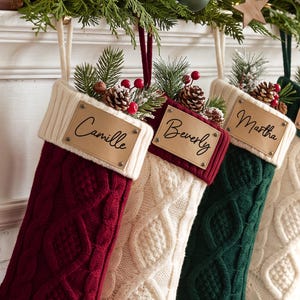 May include: Three cable-knit Christmas stockings in deep red, cream, and dark green. Each stocking features a cream-colored ribbed cuff with a rectangular tan leather patch. The patches are engraved with the names Camille, Beverly, and Martha.