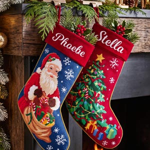 May include: Two needlepoint Christmas stockings with red velvet cuffs. The blue stocking features Santa Claus with a gift sack, while the red stocking displays a decorated Christmas tree. The names Phoebe and Stella are embroidered in white script.