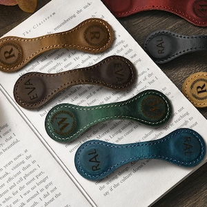 Op de afbeelding: Leren magnetische bladwijzers in diverse kleuren, elk met een gepersonaliseerde initiaal of naam. De ovale bladwijzers hebben een gestikte rand en klemmen op boekpagina's. Kleuren: bruin, groen en blauw.
