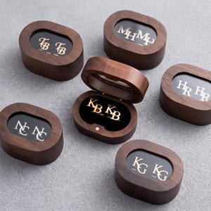 May include: Six oval-shaped dark wood boxes with hinged lids and clear glass windows. Each box features gold-colored metal initials visible through the glass, including TB, MHMH, HRHR, NCNC, KBKB, and KGKG. One box is open, revealing the gold initials inside.
