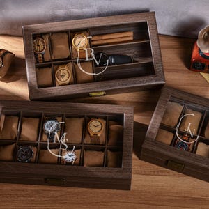 Puede incluir: Cajas de exhibici&oacute;n de relojes de madera con tapas de vidrio. Las cajas est&aacute;n llenas de varios relojes y almohadas para relojes. Una caja tiene un compartimento para puros y una llave de coche. Las cajas est&aacute;n grabadas con iniciales.