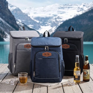 May include: Three insulated cooler backpacks in gray, navy blue, and black, each with a personalized leatherette patch. The navy blue backpack has the initials "RS", the gray backpack has "MK", and the black backpack has "SH". Two beer bottles and a glass of amber liquid are also visible.