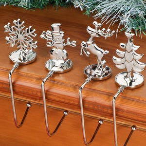 Può includere: Set di quattro porta calze natalizie argentati. Ogni supporto presenta un design festivo diverso: un fiocco di neve, un pupazzo di neve, una renna e un albero di Natale. I supporti hanno un gancio per appendere le calze e sono esposti su una superficie di legno.