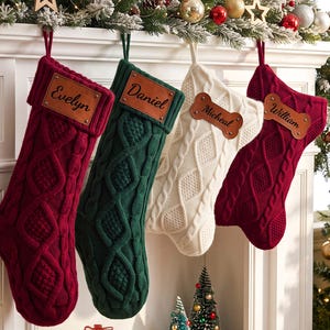 May include: Four cable-knit Christmas stockings hanging in a row. Two are deep red, one is dark green, and one is cream. Each features a personalized brown leather tag with names Evelyn, Daniel, Micheal, and William engraved in black script.