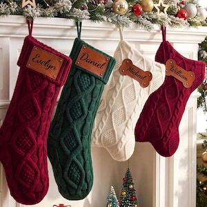 May include: Four cable-knit Christmas stockings in red, green, and cream colors. Each stocking features a hanging loop and a personalized leather-like name tag. The tags are rectangular or bone-shaped, displaying the names Evelyn, Daniel, Micheal, and William.
