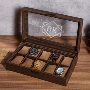 Puede incluir: Una caja de exhibici&oacute;n de relojes de madera con tapa de cristal transparente, que contiene varios relojes de pulsera. La caja es marr&oacute;n con un interior marr&oacute;n claro y un monograma en el cristal. Los relojes tienen varios dise&ntilde;os de correa y esfera.