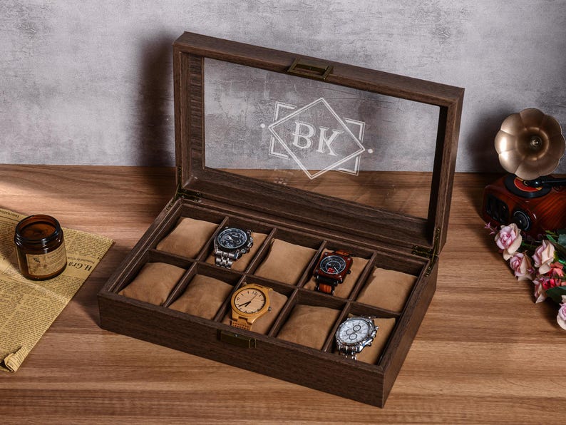 Puede incluir: Una caja de exhibici&oacute;n de relojes de madera con tapa de cristal transparente, que contiene varios relojes de pulsera. La caja es marr&oacute;n con un interior marr&oacute;n claro y un monograma en el cristal. Los relojes tienen varios dise&ntilde;os de correa y esfera.