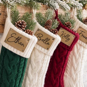 May include: A set of four cable-knit Christmas stockings in green, white, and red. Each stocking features a rectangular tan leather tag with a name printed in black script, including Ethan, Bella, Kelly, and Frank, topped with pine and pinecones.