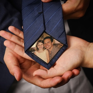 Puede incluir: Corbata azul marino con un patrón de rayas diagonales. La corbata presenta una pequeña fotografía enmarcada de un hombre y una niña. La corbata se sostiene abierta, revelando la fotografía.