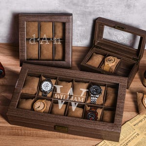 Puede incluir: Cajas de exhibici&oacute;n de relojes de madera con tapas de vidrio, que muestran varios relojes. Una caja est&aacute; abierta, revelando un reloj con una correa de color claro. Otra caja muestra las iniciales "dAn".