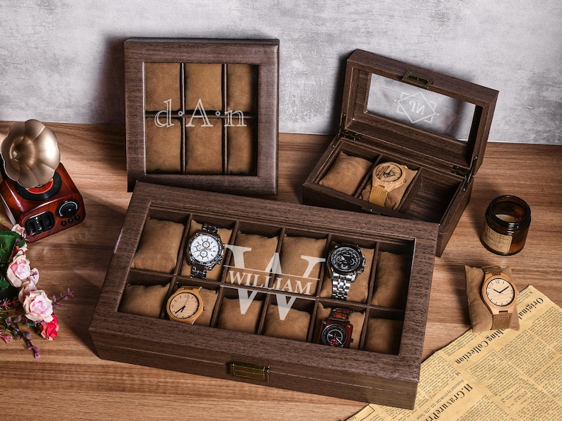 Puede incluir: Cajas de exhibici&oacute;n de relojes de madera con tapas de vidrio, que muestran varios relojes. Una caja est&aacute; abierta, revelando un reloj con una correa de color claro. Otra caja muestra las iniciales "dAn".