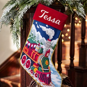 May include: A Christmas stocking featuring a needlepoint design of a colorful steam train traveling through a snowy mountain landscape. The top cuff is solid red velvet with the name Tessa embroidered in white script. Gold piping edges the stocking.
