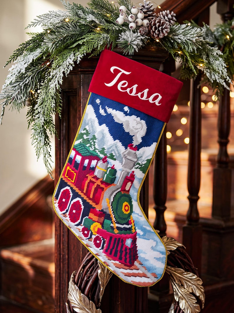 May include: A Christmas stocking featuring a needlepoint design of a colorful steam train traveling through a snowy mountain landscape. The top cuff is solid red velvet with the name Tessa embroidered in white script. Gold piping edges the stocking.